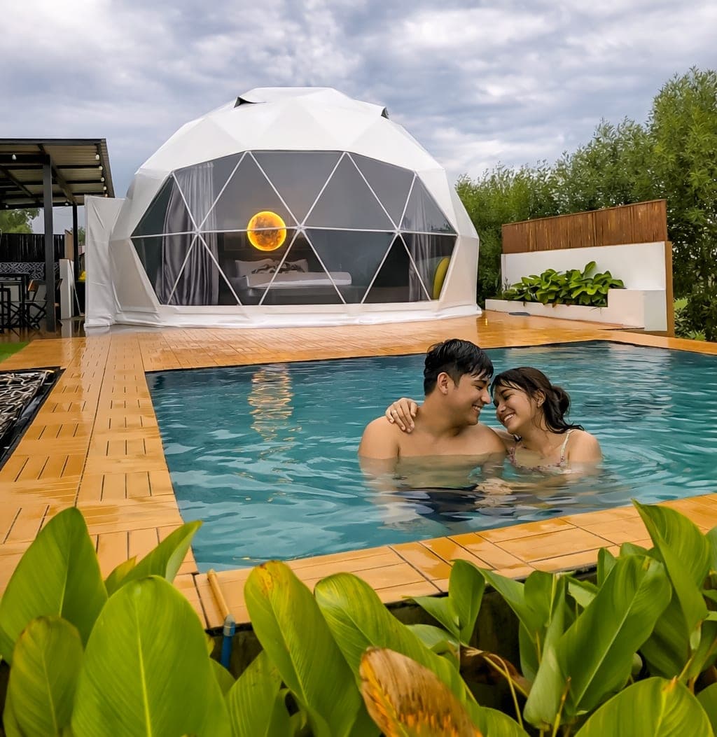 A couple in a private pool, heads together, with a lit glamping dome behind them